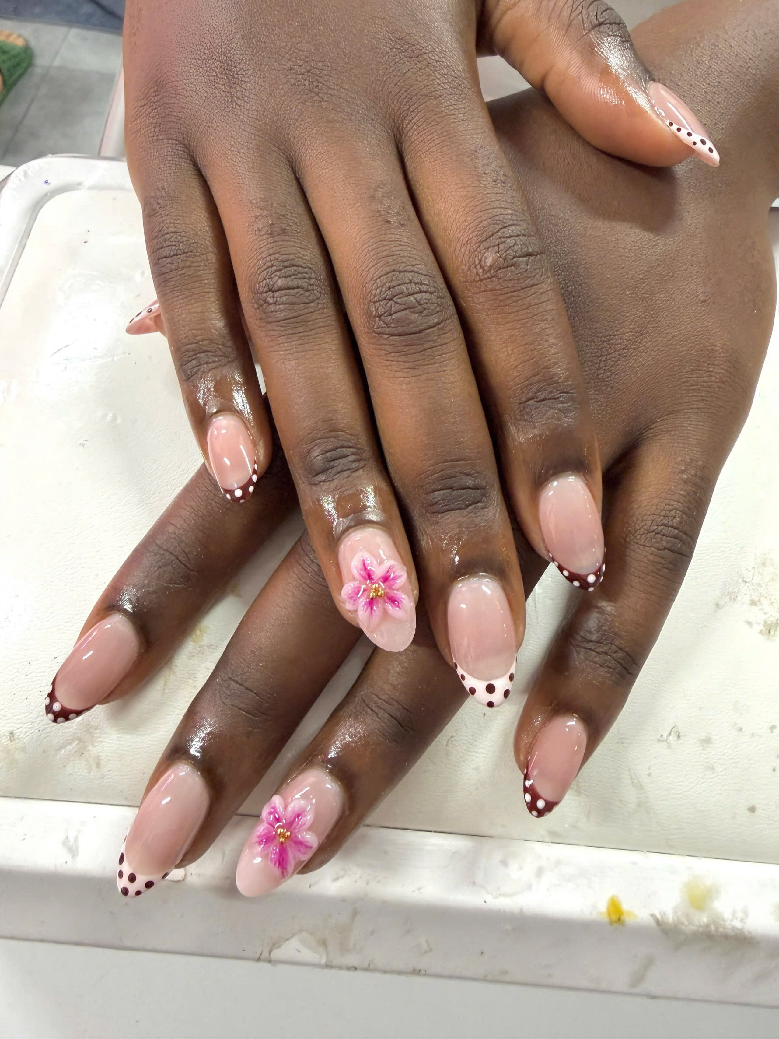Two hands with medium almond-shaped glossy nude nails, dark brown dotted French tips, and pink floral nail art with gold centers