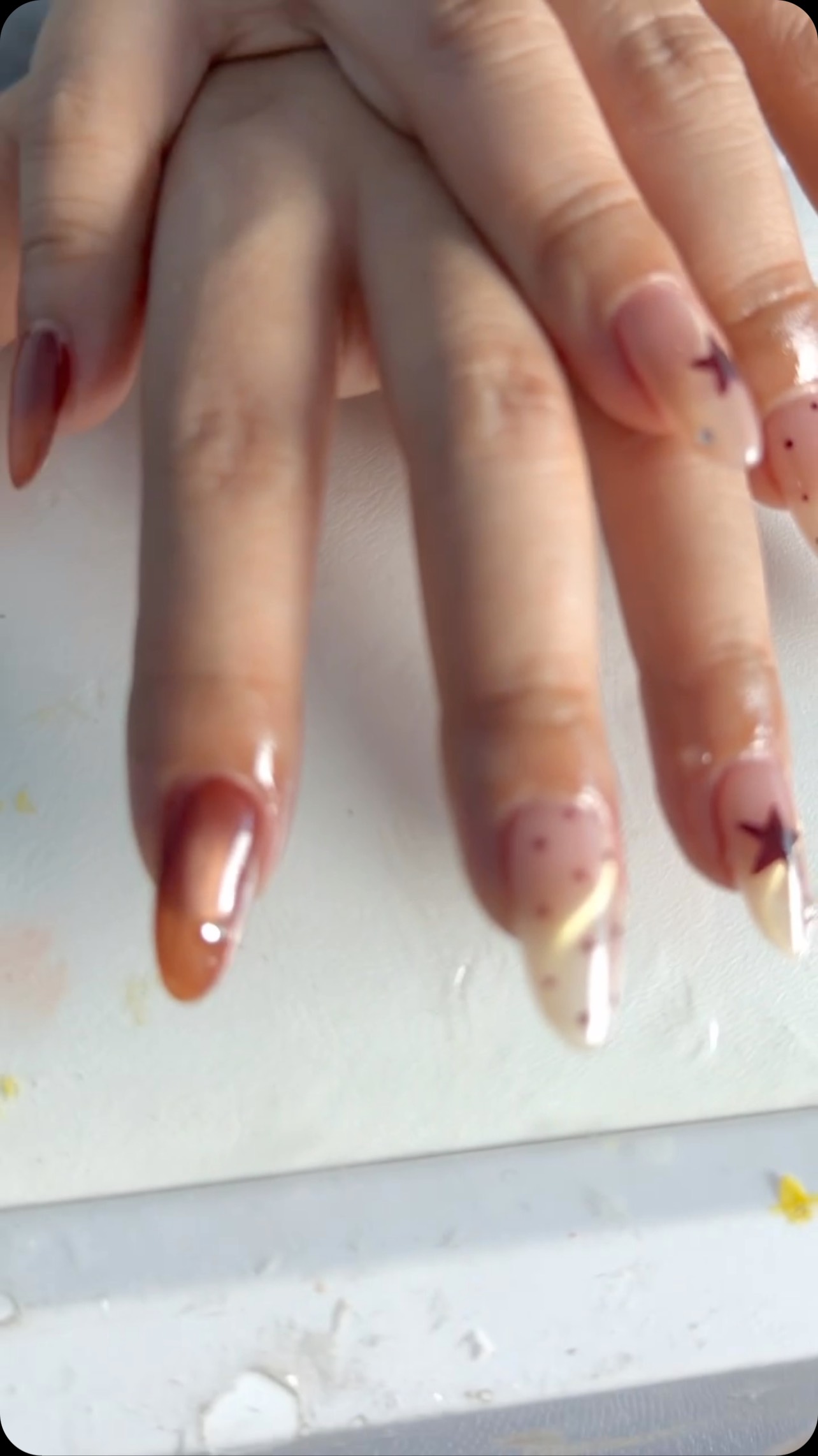Close-up of a hand with long almond-shaped nails in glossy nude and brown polish with star details, dots, and French tips