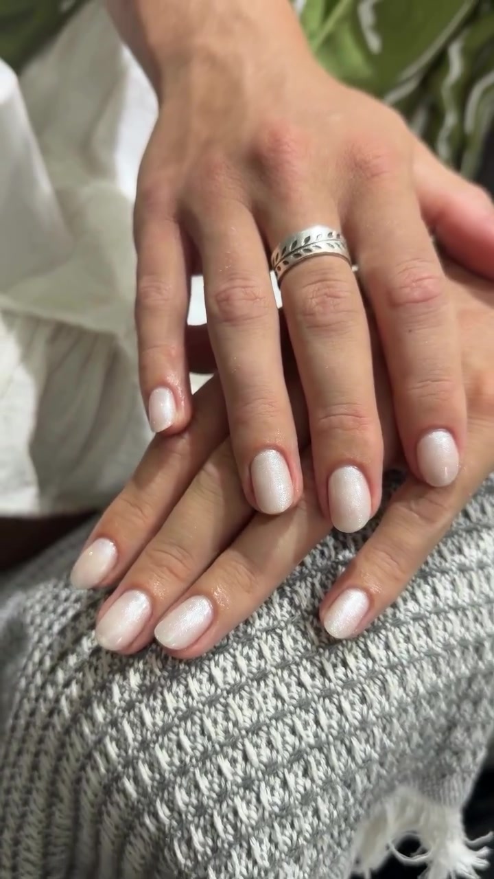 Two hands resting on a textured gray surface with short oval nails in a glossy pearlescent white and pink finish