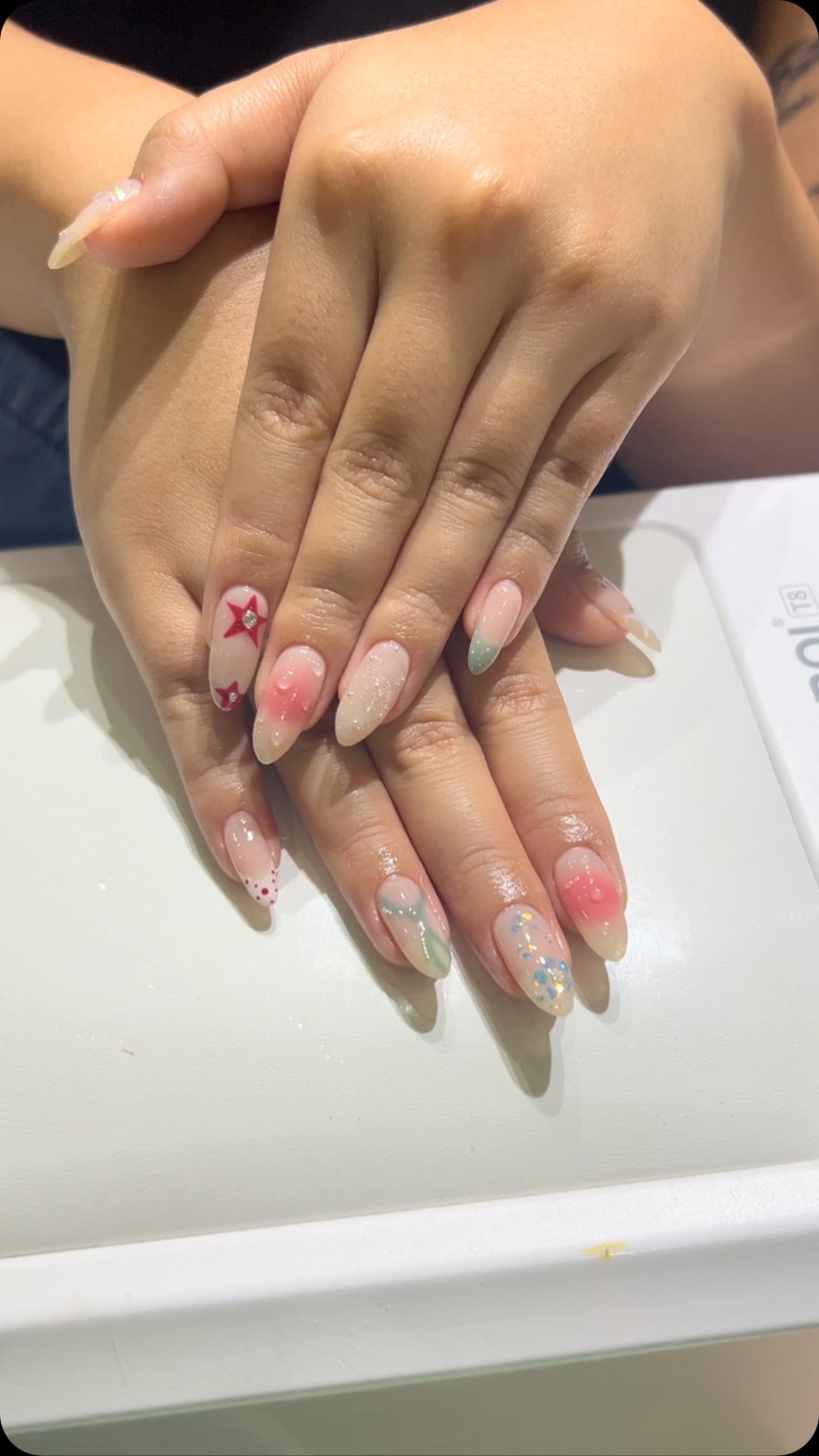 Two hands with medium oval nails featuring a sheer nude-pink base, a red star accent with a rhinestone, dotted tips, and scattered pastel glitter and small floral details.