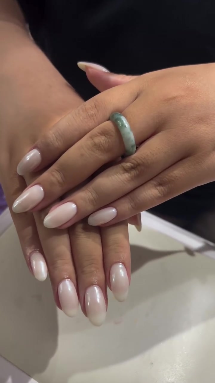 Two hands with medium almond-shaped nails in a glossy pearlescent pink-to-white chrome finish, with a green ring on one finger