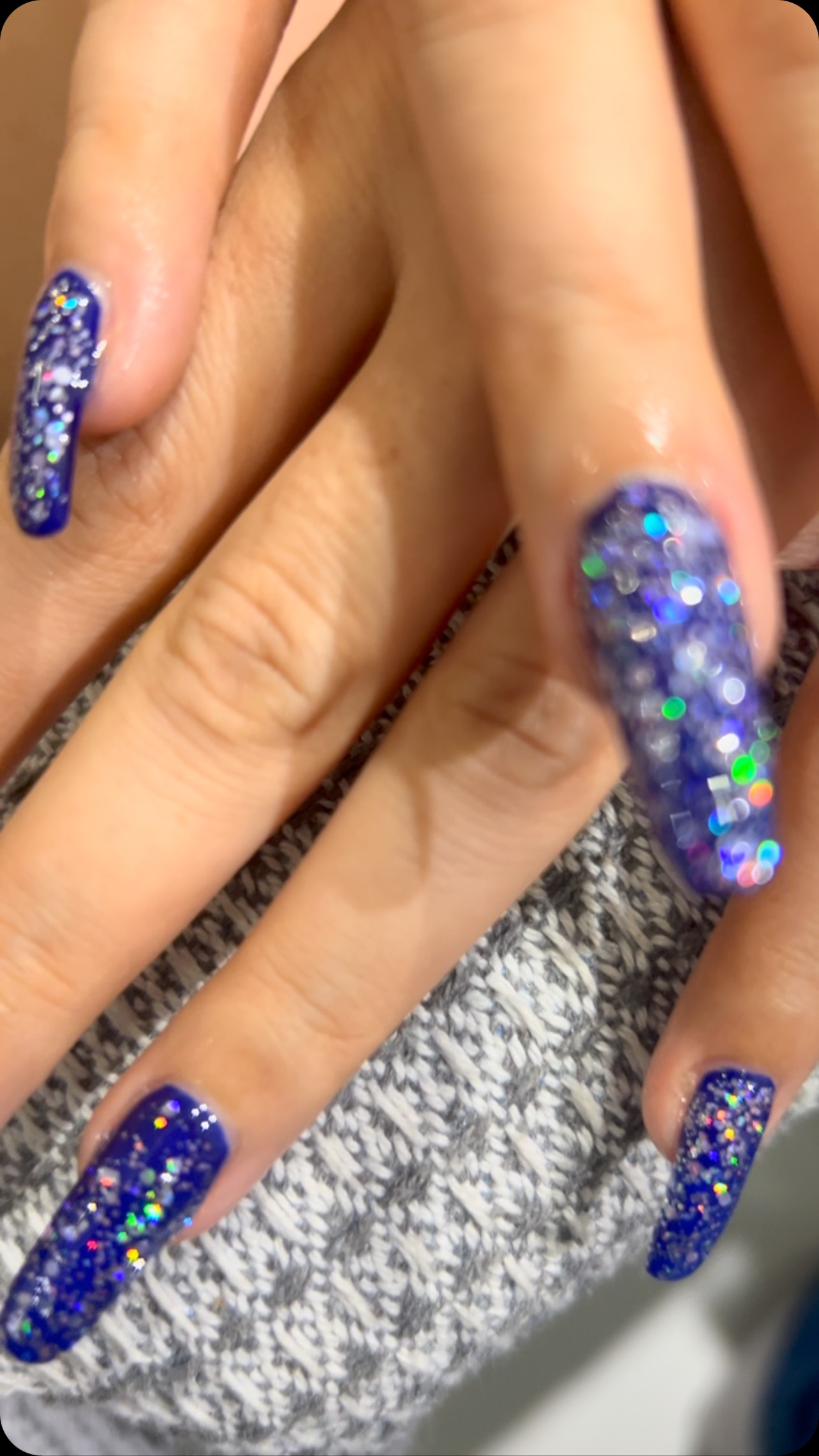 Close-up of a hand with long coffin-shaped blue and purple glitter nails resting on textured gray fabric