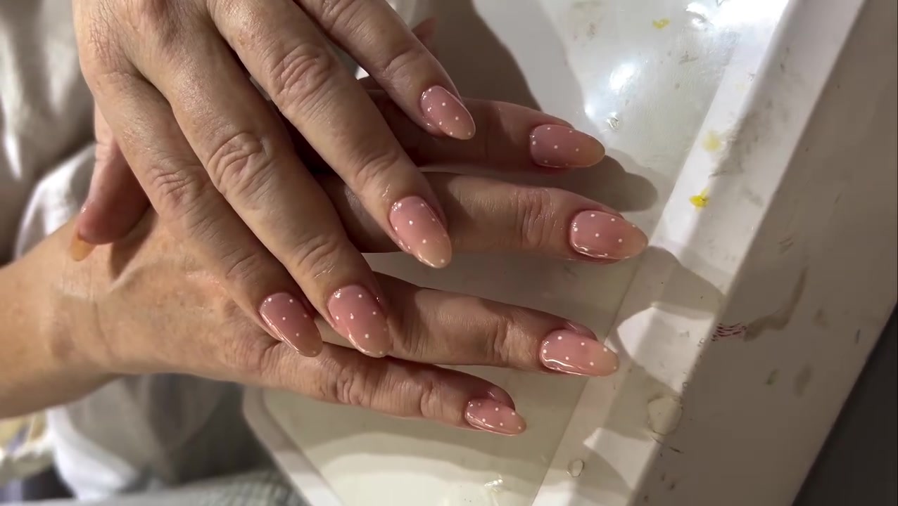 Two hands with glossy light pink oval nails decorated with small white dots resting near a white surface