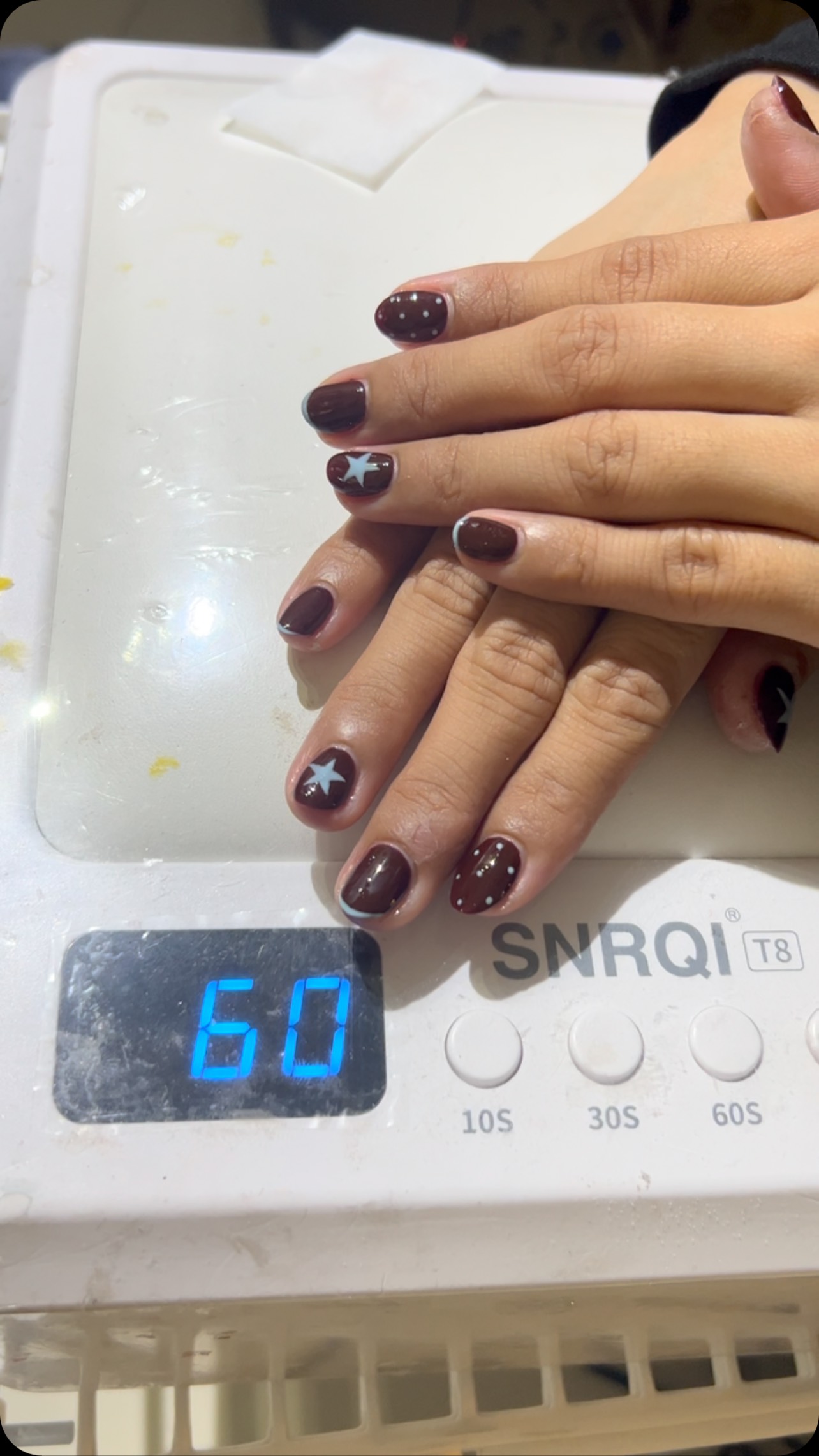 Two hands with short square nails in glossy dark brown polish decorated with white stars and dotted accents