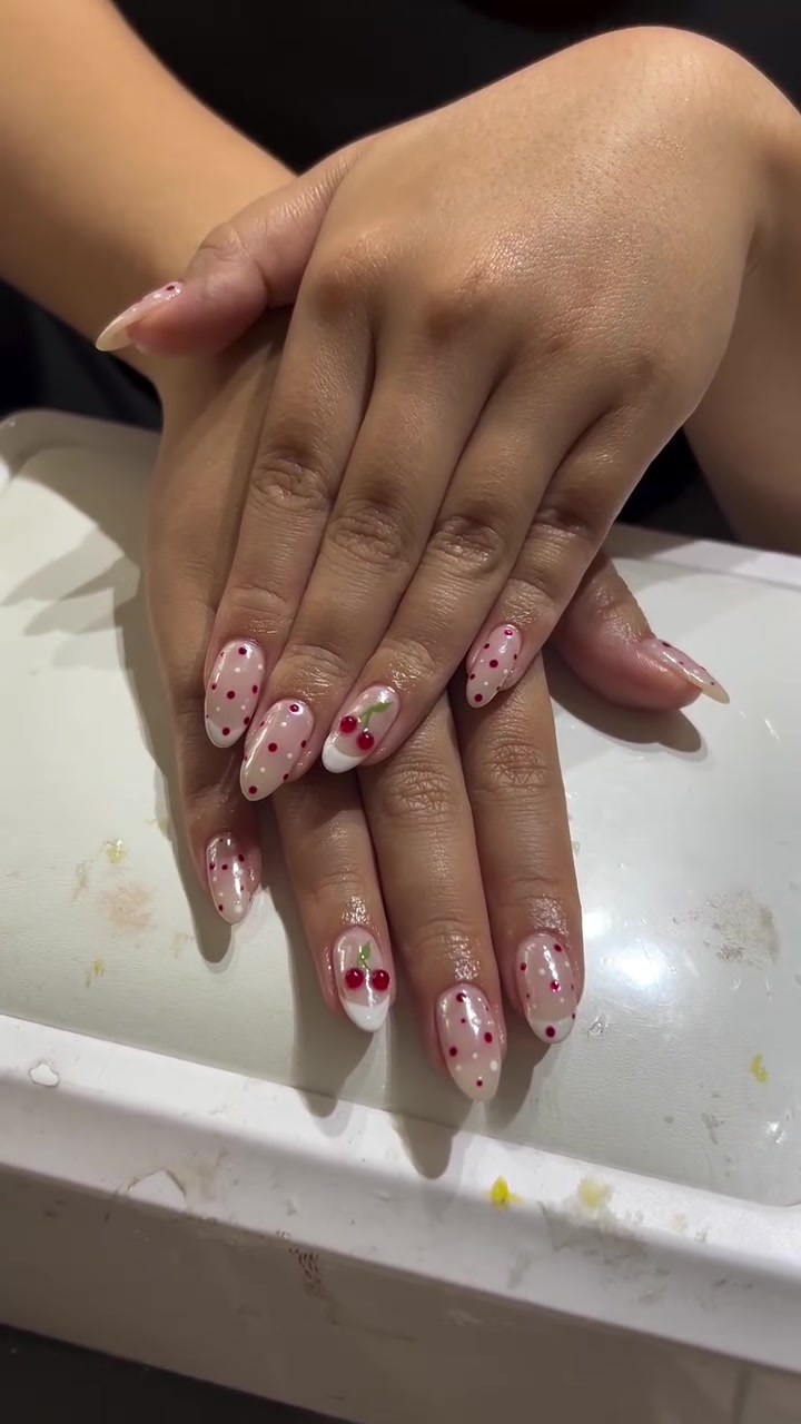 Two hands with medium almond-shaped nails in a sheer pink base, white tips, red polka dots and cherry nail art