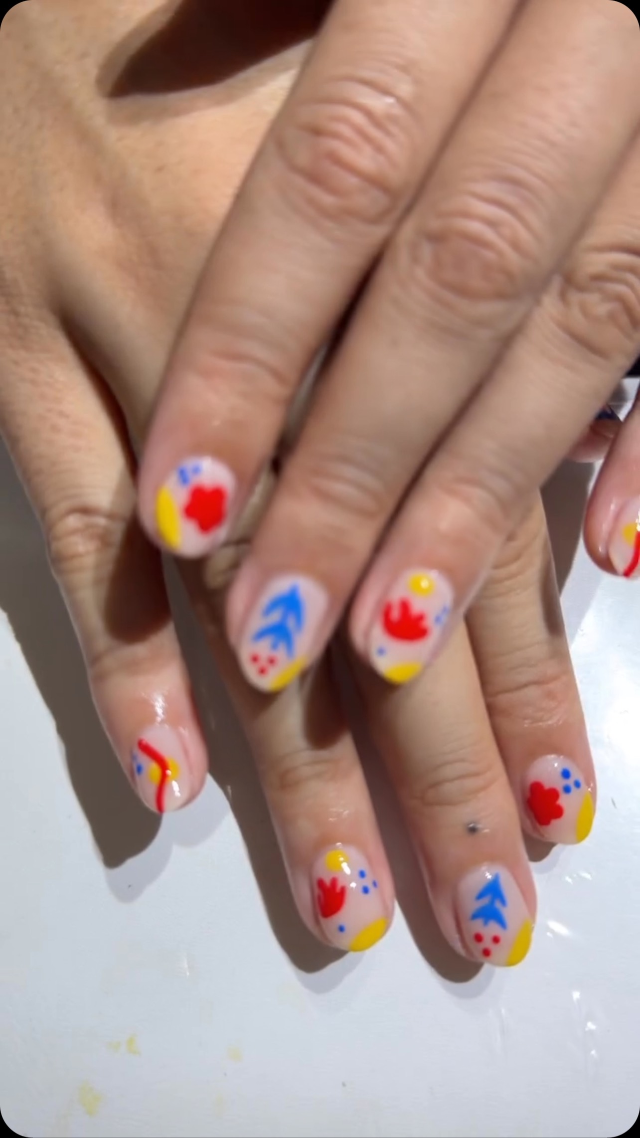 Close-up of two hands with glossy medium-length oval nails in a colorful abstract design with pink bases and red, blue and yellow shapes, dots and lines