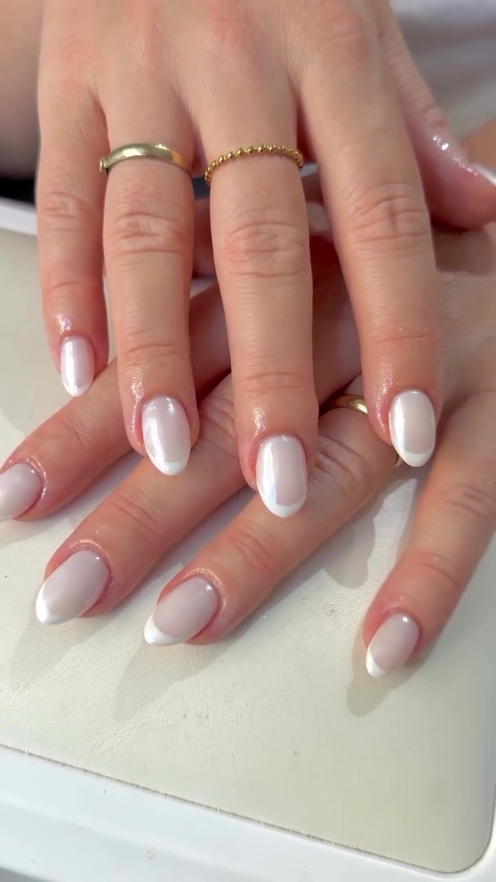 Close-up of two hands with medium oval nails showing glossy white French tips over a sheer pink base, with two gold rings visible