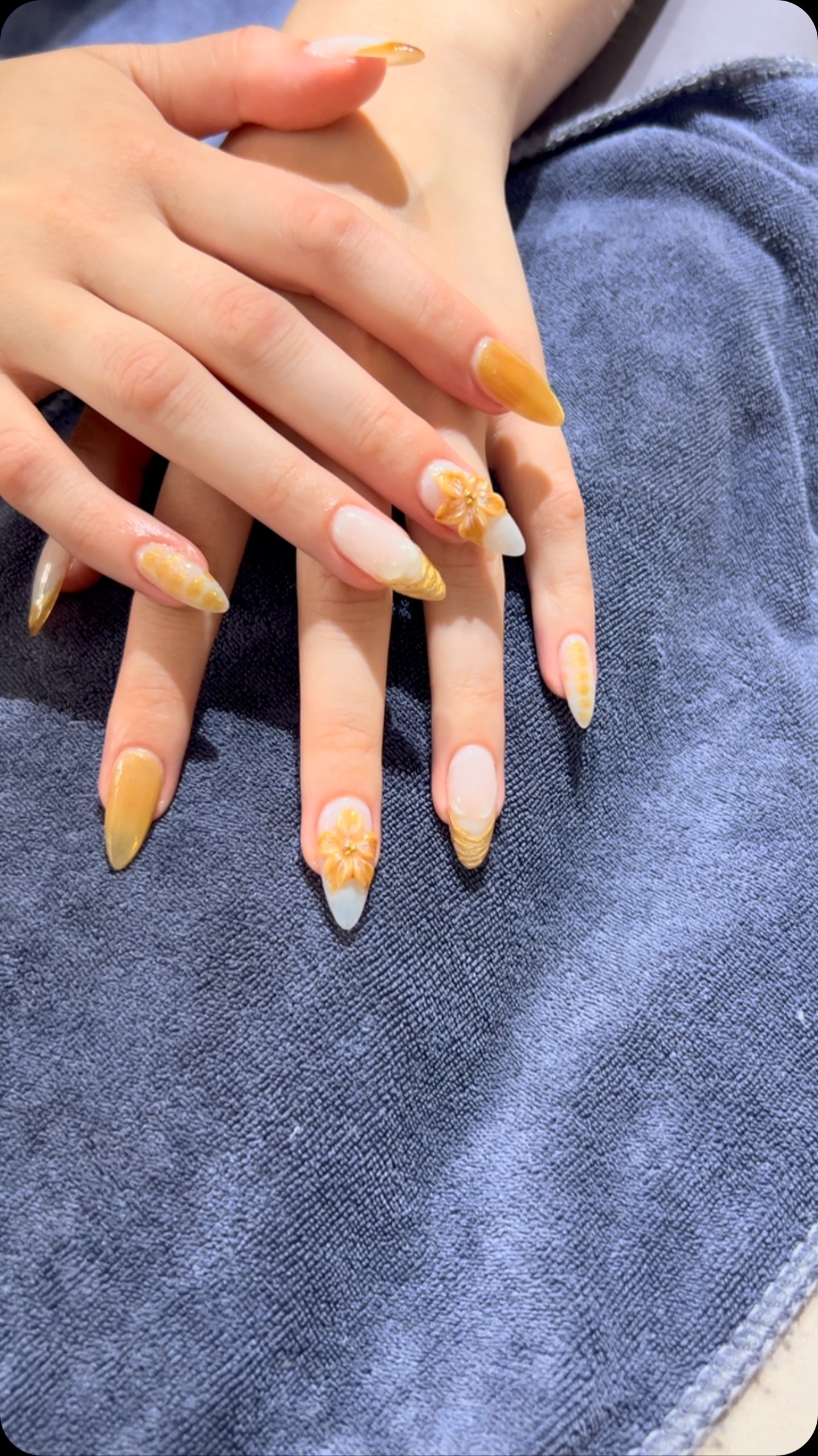 Two hands with long almond-shaped nails in glossy yellow and white designs, with gold floral accents and glittery tips, resting on blue fabric