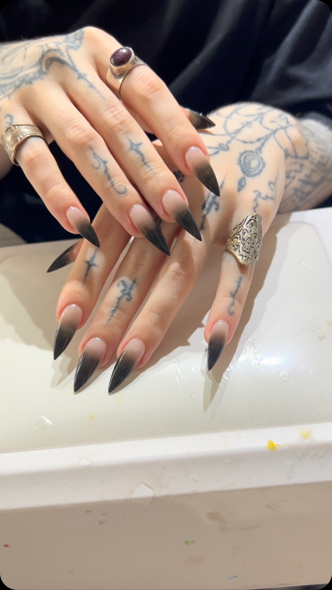 Close-up of a hand with long stiletto nails in a black-to-nude ombre gradient with a glossy finish, worn with rings