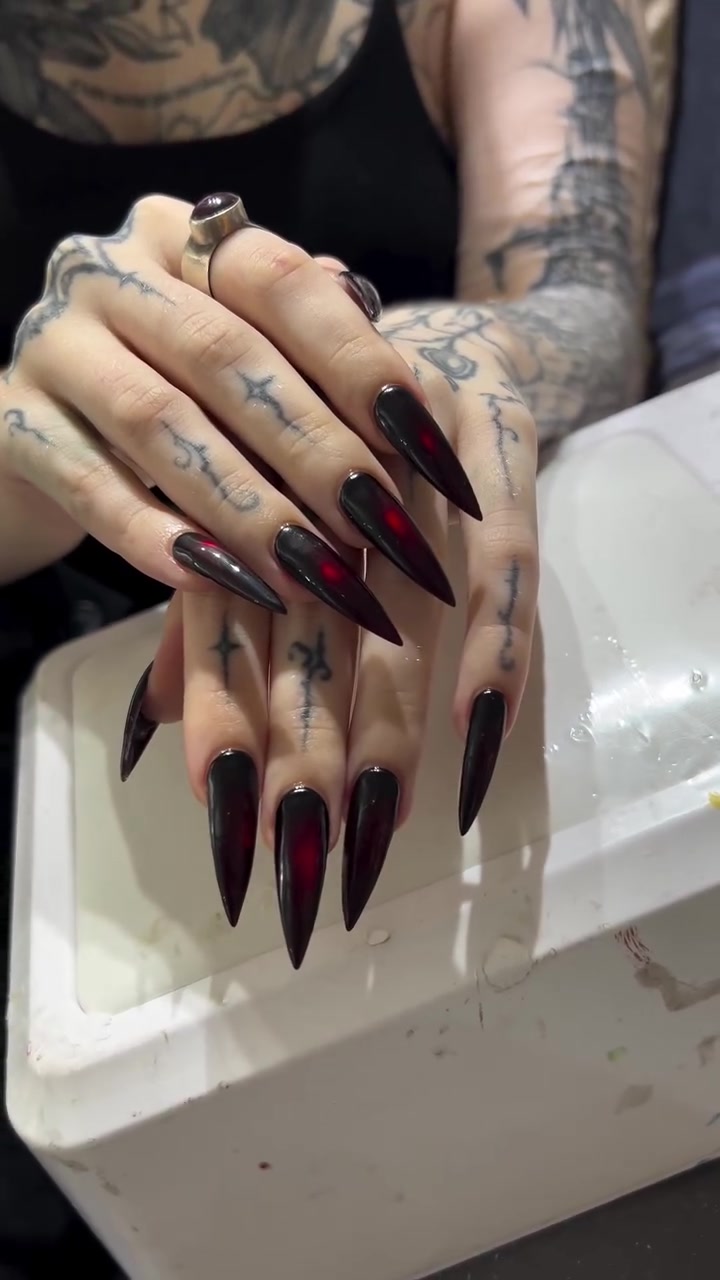 Two hands with long glossy black stiletto nails featuring red centers, resting on a white surface