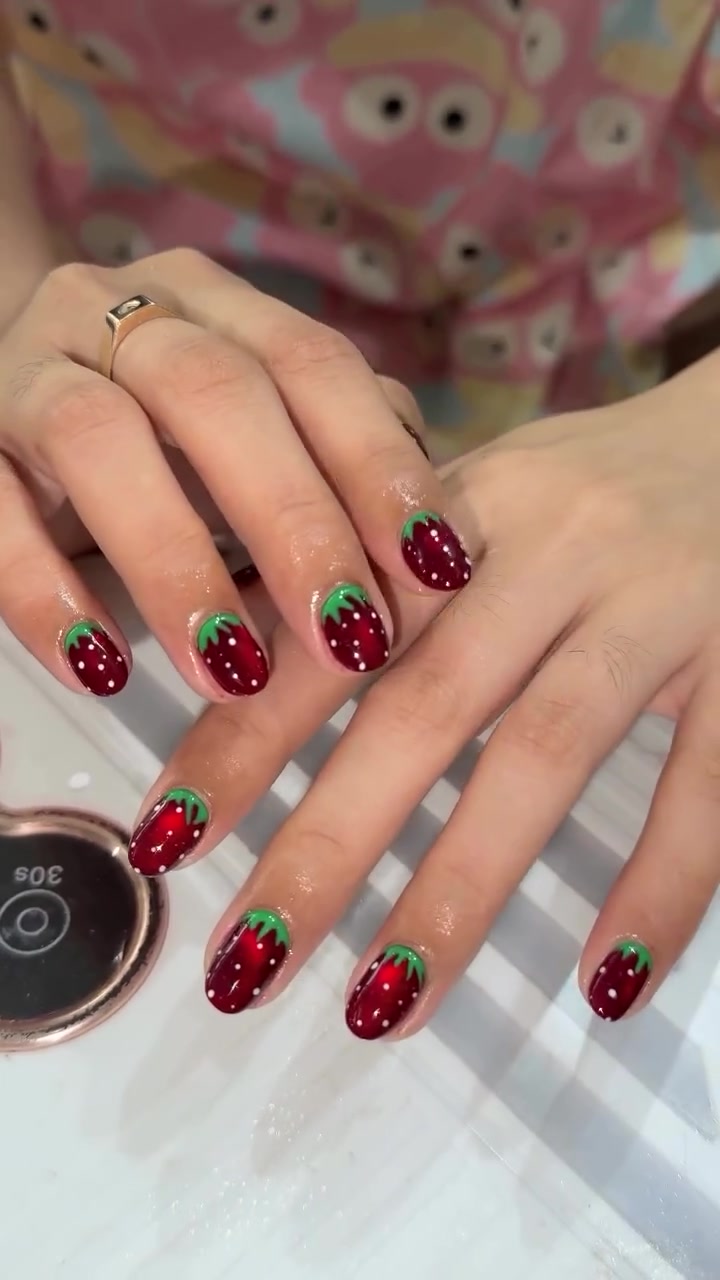 Close-up of hands with medium almond nails painted with a glossy red base, green tips, and small white dots like strawberries